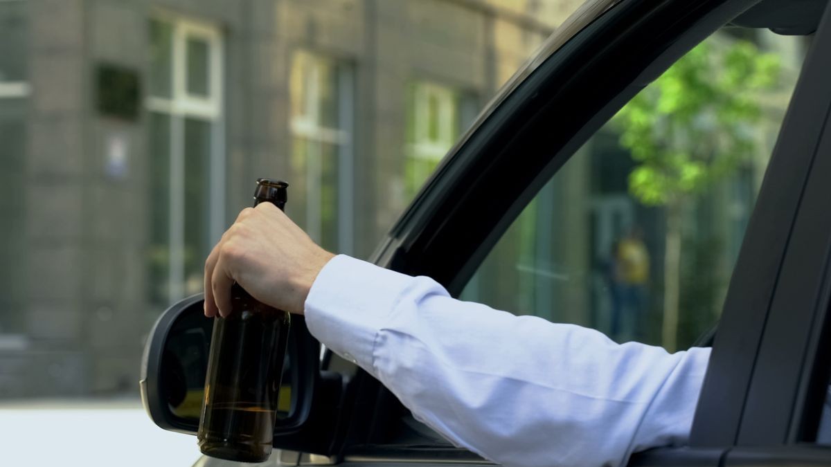 Man holding beer bottle inside car, drinking alcohol while driving, crash risk Man holding beer bottle inside car, drinking alcohol while driving, crash risk